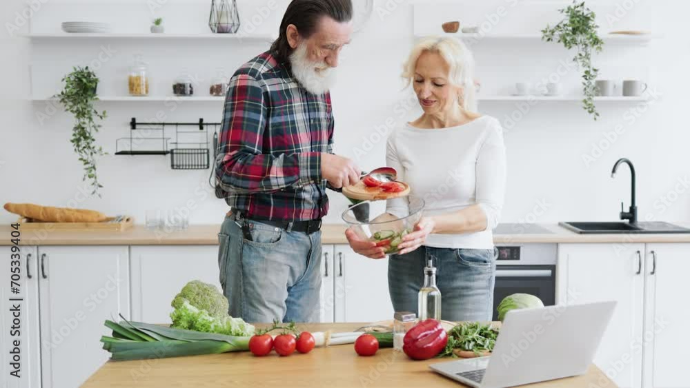 Charming elderly couple husband and wife spend free time together in kitchen making healthy salad from fresh vegetables. Man with gray beard pours chopped pieces of tomato into bowl held by partner. Charming elderly couple husband and wife spend free time together in kitchen making healthy salad from fresh vegetables. Man with gray beard pours chopped pieces of tomato into bowl held by partner.