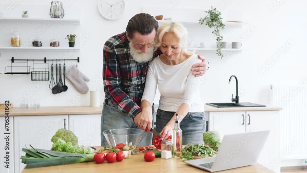Positive retired couple cutting pepper while holding knife together in hands and putting pieces into glass bowl. Old family spends leisure time making healthy salad in modern kitchen. Positive retired couple cutting pepper while holding knife together in hands and putting pieces into glass bowl. Old family spends leisure time making healthy salad in modern kitchen.