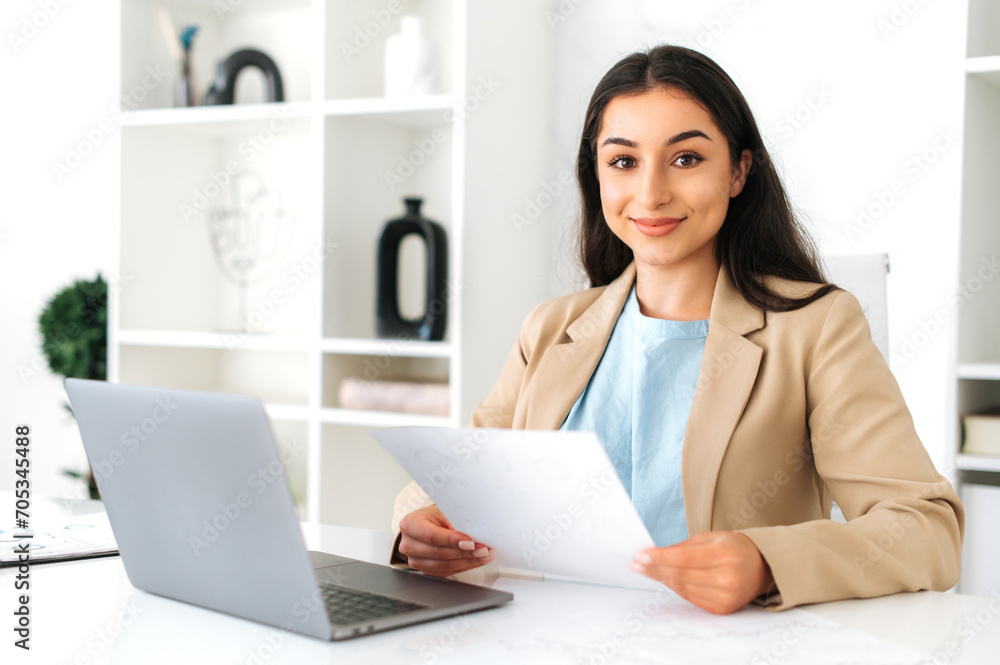 Confident successful brunette indian or arabian business woman in an elegant suit, ceo, company employee, sitting at a work desk in the modern office, holds documents, looking at camera and smiling Confident successful brunette indian or arabian business woman in an elegant suit, ceo, company employee, sitting at a work desk in the modern office, holds documents, looking at camera and smiling