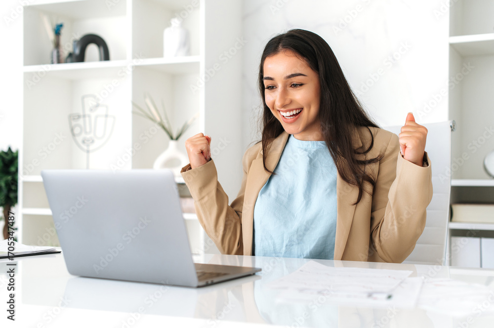 Joyful happy indian or arabian business woman, sitting at a work desk in the office, happily looks at laptop screen, rejoicing promotion at work, big profit, good work results, gesturing hands Joyful happy indian or arabian business woman, sitting at a work desk in the office, happily looks at laptop screen, rejoicing promotion at work, big profit, good work results, gesturing hands