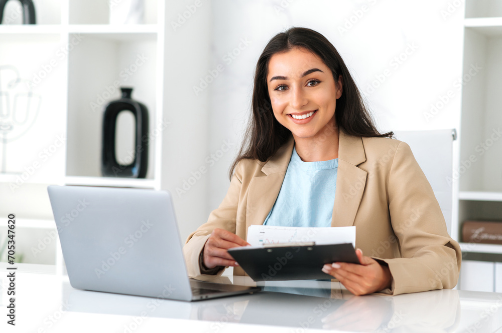 Beautiful positive successful indian or arabian business woman, financial manager sits at a work desk in the office, working on a laptop, studying financial reports, glad with results,smiles at camera Beautiful positive successful indian or arabian business woman, financial manager sits at a work desk in the office, working on a laptop, studying financial reports, glad with results,smiles at camera