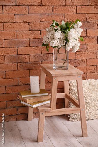 Vase with lilac flowers, candle and books on stepladder near brick wall in room
