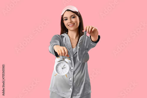 Young woman in pajamas with alarm clock and sleeping mask on pink background