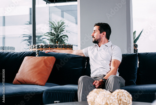 man sitting on a sofa looking to the side, in a living room wearing a T-shirt and slacks
