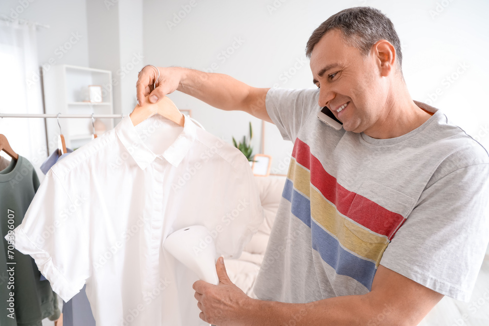 Obraz premium Mature man steaming clothes while talking by mobile phone at home