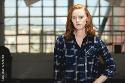 Young red haired woman in plaid shirt stands in an industrial space.
