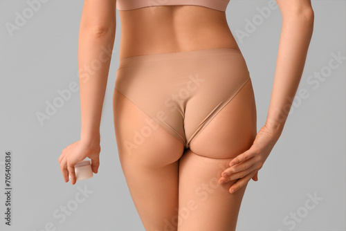 Beautiful young woman with jar of anti-cellulite cream on grey background, back view