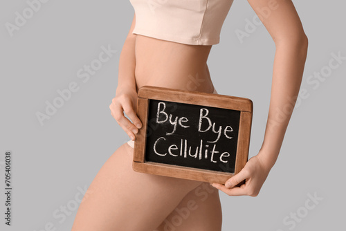 Beautiful young woman holding board with text BYE BYE CELLULITE on grey background