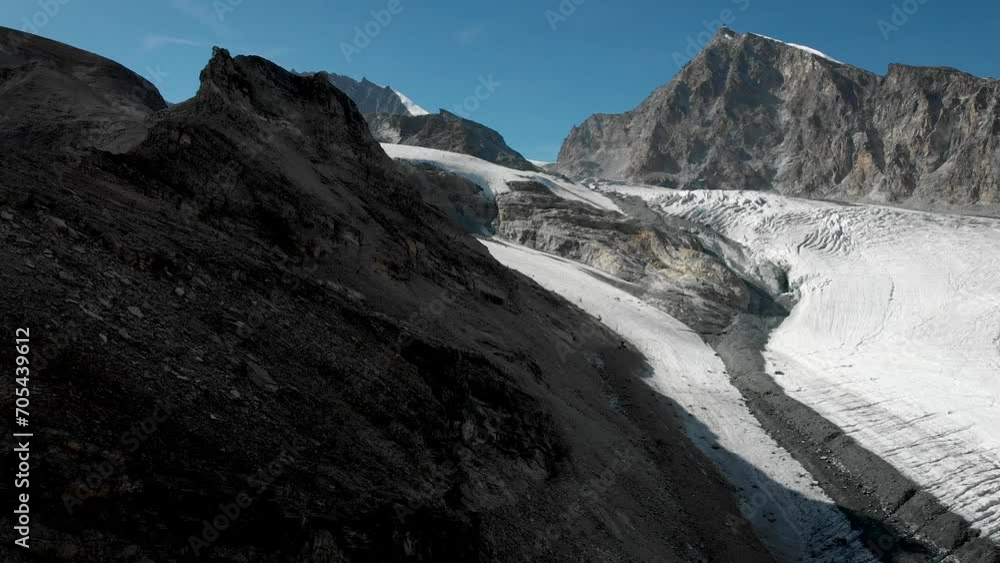 Aerial flyover alongside a cliff next to the Allalin glacier with Allalinhorn peak in view near Saas-Fee in Valais, Switzerland on a sunny summer day in the Swiss Alps.