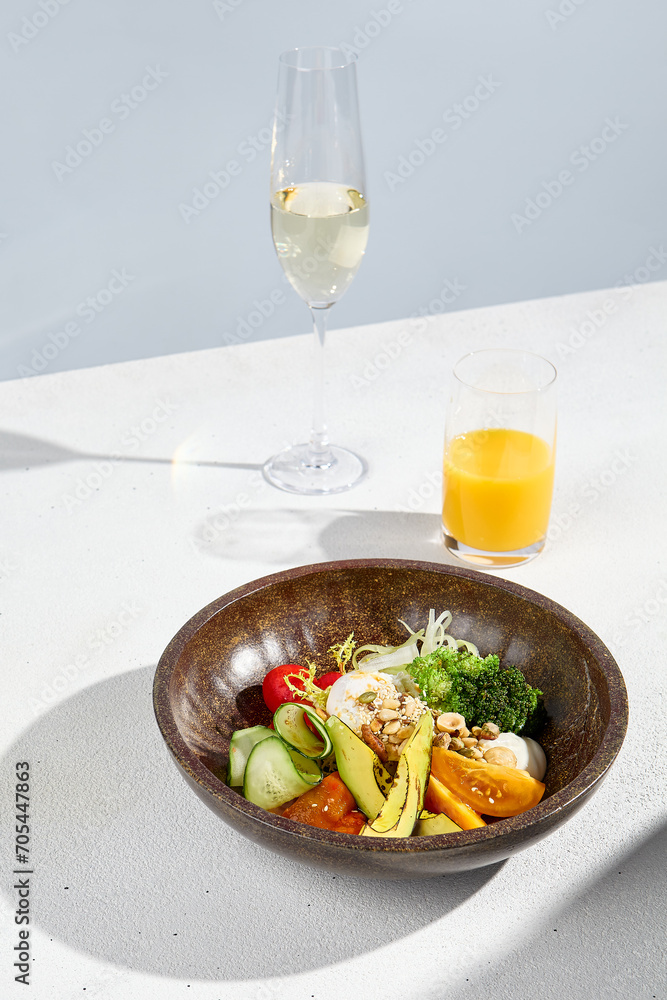 Salad bowl with avocado, vegetables, poached egg, with orange juice and sparkling wine in hard sunlight
