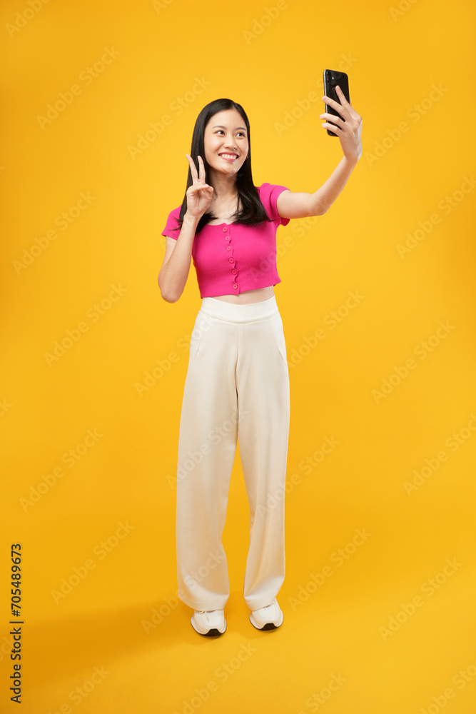 Portrait of a young Asian woman taking a selfie photo on a smartphone with a joyful expression in casual clothing and looking at a camera. Isolated on a yellow background. Portrait of a young Asian woman taking a selfie photo on a smartphone with a joyful expression in casual clothing and looking at a camera. Isolated on a yellow background.