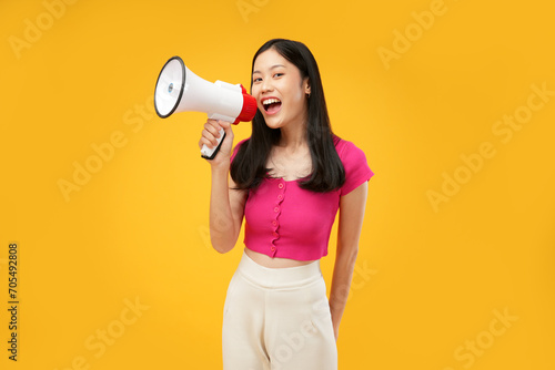 Photo of a young Asian woman wearing a pink t-shirt, holding a megaphone with a Joyful expression. Isolated on a yellow background. 