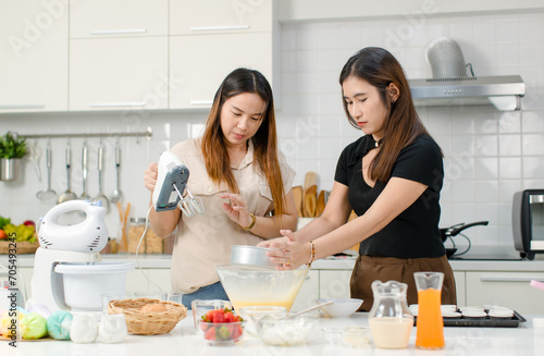 Asian cheerful professional female pastry bakery baker chef housewife with friend standing smiling helping sifting flour into glass bowl with stainless sifter preparing dough in decorated kitchen