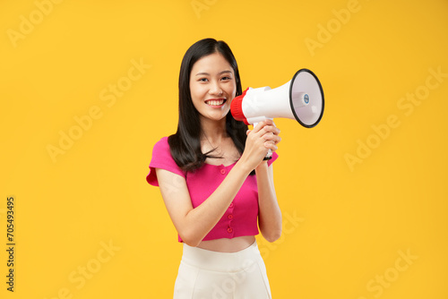Portrait of a young Asian woman holding a megaphone in her hands and looking at the camera. Wearing a pink t-shirt and an isolated yellow background.