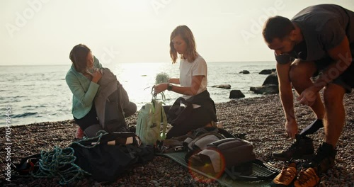 Two blonde girls and a brunette guy in a gray T-shirt are dismantling their backpacks and preparing ammunition to create a belay while starting their rock climbing activity on a rocky seashore in photo