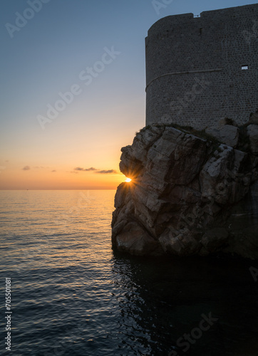 The setting sun between rocks near the ocean and walls of Dubrovnik