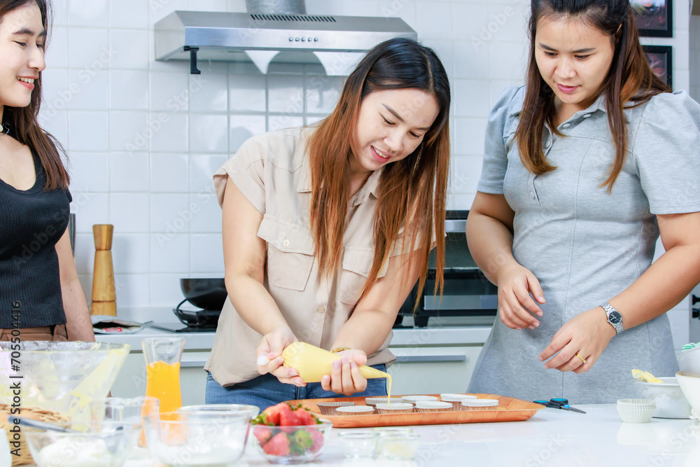 Asian happy professional female pastry bakery baker chef housewife with close friends standing smiling helping using tube bag squeezing whipped cream decorating topping preparing cupcakes in kitchen