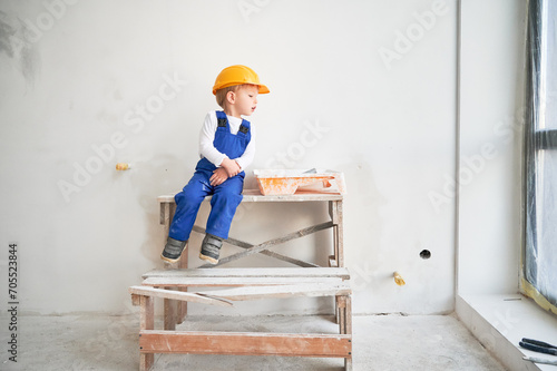 Cute kid construction worker sitting on wooden table against white wall in apartment under renovation. Cheerful little boy wearing safety helmet and work overalls while playing at home.