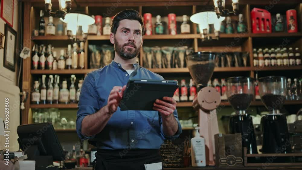 Competent bartender with welcoming smile on face working on digital tablet while standing at bar counter. Caucasian man in blue shirt and black apron preparing for working shift with device in hands. Competent bartender with welcoming smile on face working on digital tablet while standing at bar counter. Caucasian man in blue shirt and black apron preparing for working shift with device in hands.