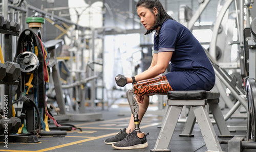 Woman with prosthetic leg sitting in gym lifting dumbbell weight. Female with foot prosthesis physical workout exercise in fitness. Artificial limb equipment help accident survivor amputee to mobility © Nassorn