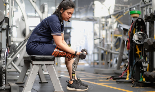 Woman with prosthetic leg sitting in gym lifting dumbbell weight. Female with foot prosthesis physical workout exercise in fitness. Artificial limb equipment help accident survivor amputee to mobility