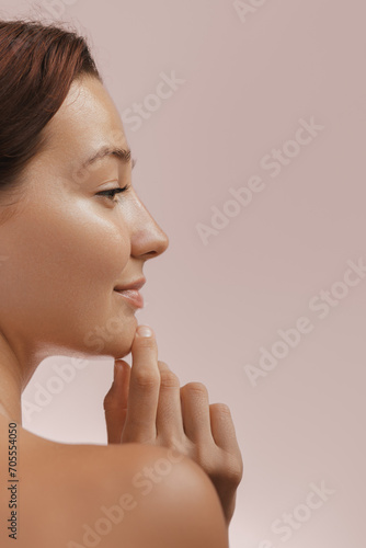 Beautiful young adult woman in profile touching her perfect face with healthy shiny skin, hand on chin. Photo of gorgeous girl after morning beauty cosmetic routine finishes daily makeup on pink
