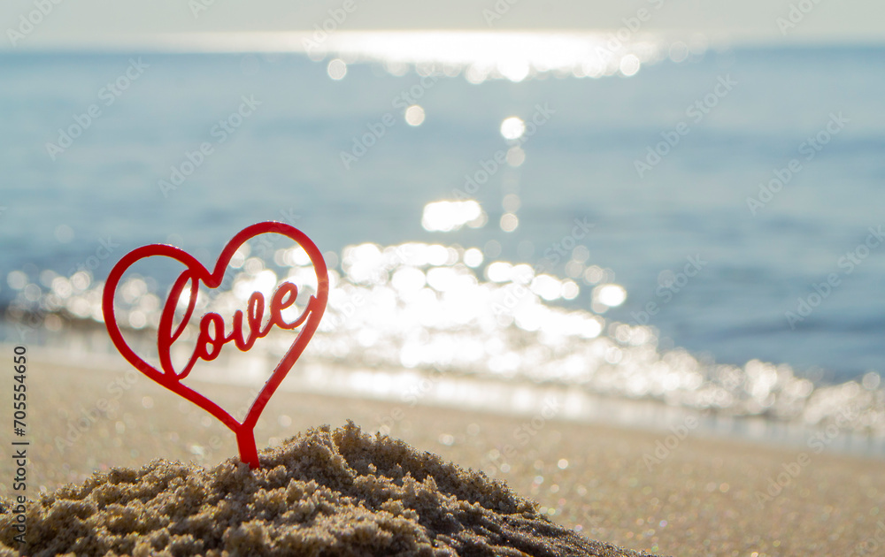 Fototapeta premium Plastic stick in shape of red heart and word Love in sand on beach seashore on sunny summer day close-up. Figures in shape of heart word Love on background sea waves. Love relationship romance concept