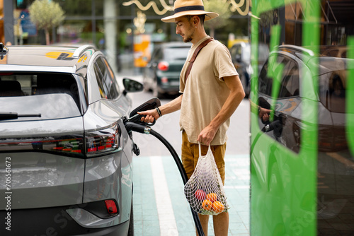 Man with groceries waiting for electric car to be charged on public charging station near a shopping mall. Man holds mesh bag with fresh vegetables. Sustainable lifestyle concept photo