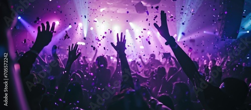 Photo of partygoers dancing under purple lights with confetti and raised hands at a shiny, crowded nightclub event.