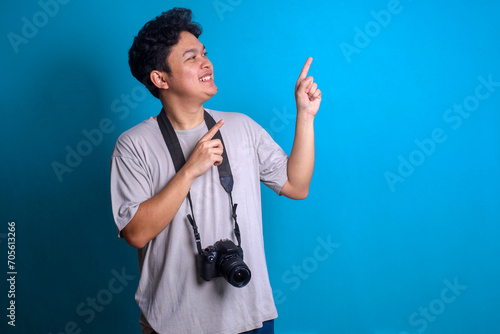 Excited young photographer bring camera and pointing finger to the side on blue background