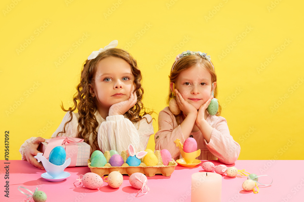 Naklejka premium Little cute girls sitting at table with panted, decorated, Easter eggs against yellow background. Concept of Easter holiday, celebration, traditions, childhood, happiness