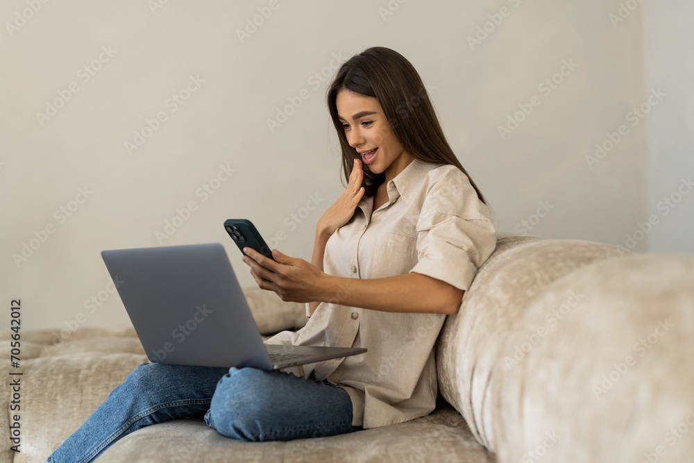 Surprised woman using phone and laptop sitting on a couch in the living room in the night at home Surprised woman using phone and laptop sitting on a couch in the living room in the night at home