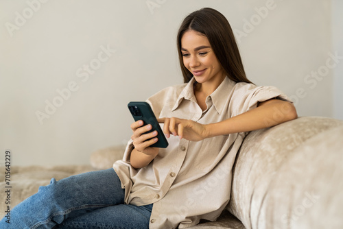 Smiling young woman relaxing on sofa with laptop holds smartphone enjoy at home