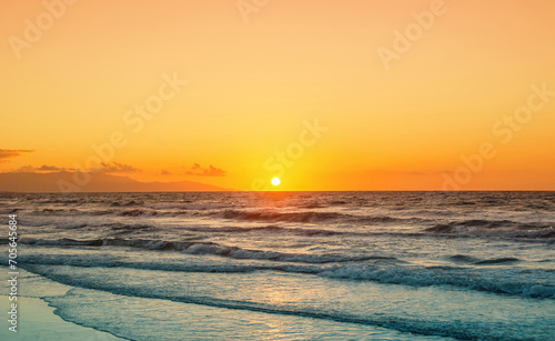 Sunset over sea, image is nearly split in half with orange sky and blue-ish water. Empty beach on sunset, mountains in the background. 