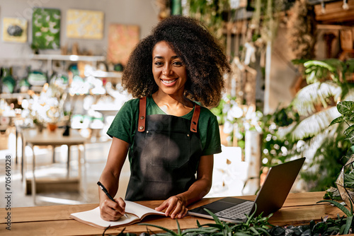 Confident successful sincere smiling charming business woman florist gardener entrepreneur in black apron and green t-shirt in plant shop writing in notebook working on laptop. photo