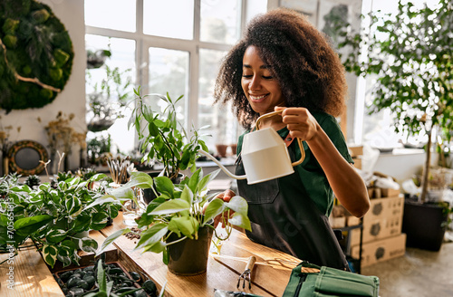 Tender care for plants. Beautiful african american woman standing by wooden table and watering green flowers in pots. Female store florist using gardening skills and knowledge at favorite work. photo