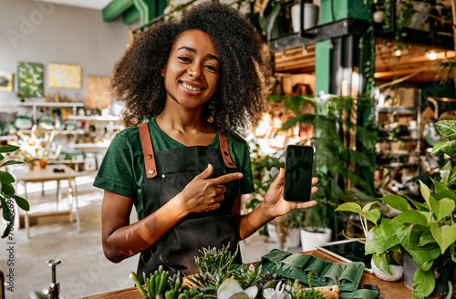 Beautiful smiling successful business lady florist holding phone and pointing finger at it while standing behind shop counter with plants and flowers. Online sales, application, order, delivery. photo