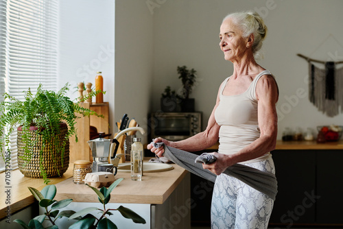 Healthy female boomer in activewear exercising with towel while standing by kitchen counter surrounded by green domestic plants photo