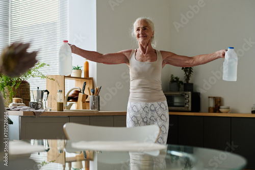 Smiling senior woman in activewear outstretching her arms and holding two plastic canisters with water while exercising in the kitchen photo