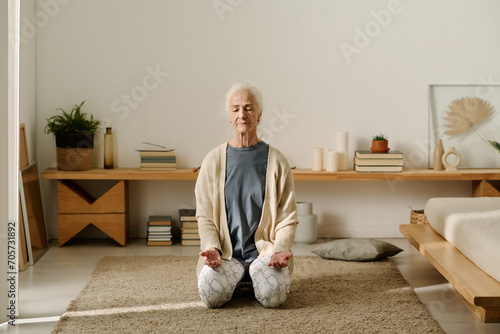 Senior woman in casualwear sitting on her knees on the floor of bedroom and keeping eyes closed while practicing yoga exercise