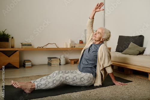 Retired woman in activewear sitting on mat on the floor of bedroom and doing side bends with raised arm while working out after sleep photo