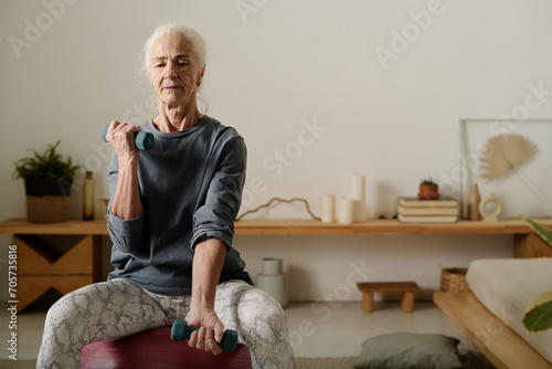 Aged woman in activewear sitting on fitball and practicing physical exercise for arm muscles with dumbbells in front of camera