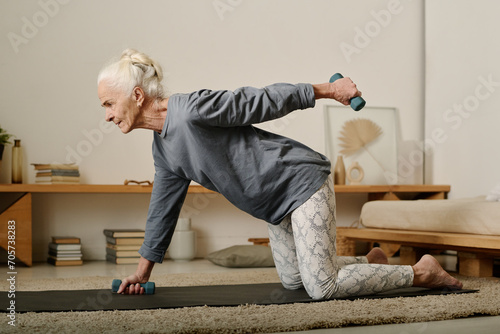 Side view of senior active woman in leggins and pullover standing on knees on the floor of bedroom and doing exercise with dumbbells photo