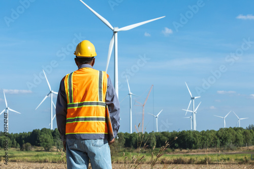 Male engineer wearing uniform safety vest and yellow helmet standing backwards looking at wind turbines farm.