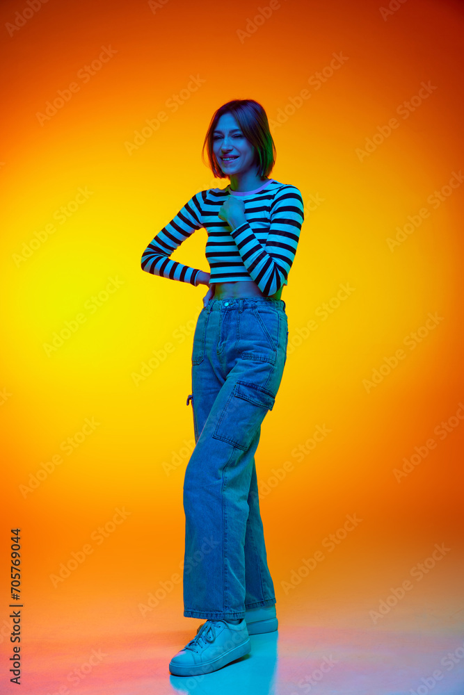 Fototapeta premium Full-length portrait of beautiful young woman dressed casual clothes and positively looking at camera against gradient orange background. Concept of beauty, youth, art, emotions, facial expression. Ad