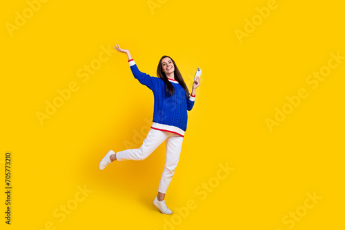 Full body length photo of hispanic woman wearing blue knit sweater raise hand above head with phone isolated on yellow color background
