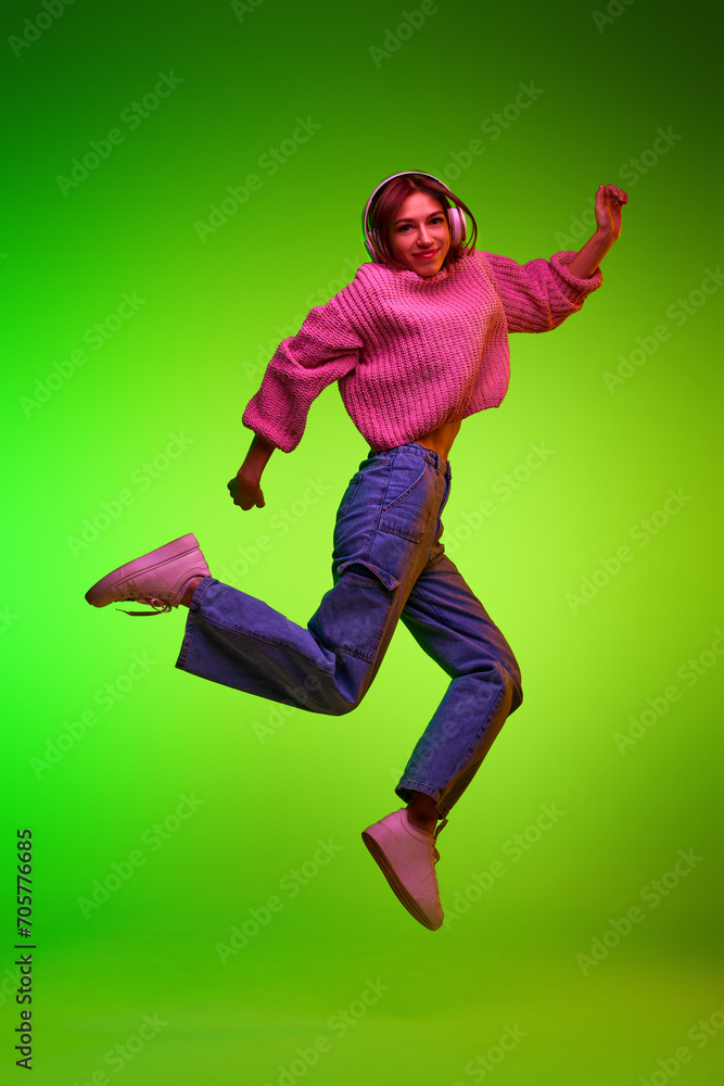 Full length portrait of young fashion dressed woman jumping in action while listening music in headphones against gradient green studio background. Concept of music and dance, technology, culture. Ad