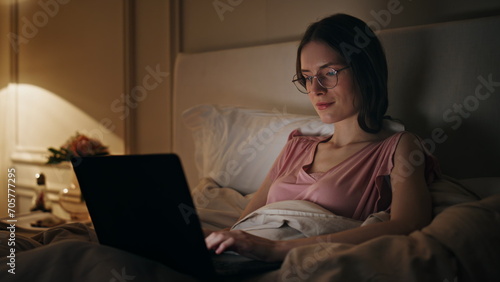 Evening girl typing laptop working in bed. Smiling relaxed woman browsing web