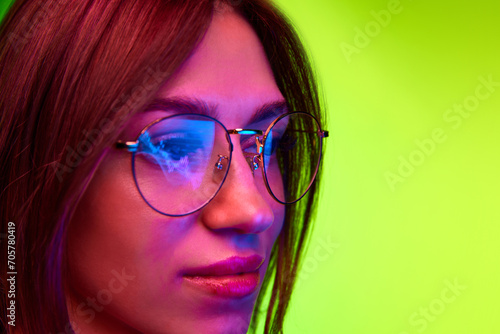 Fotografie Young business woman wearing glasses looking at computer screen with trading charts reflecting in eyeglasses against green background with copy space