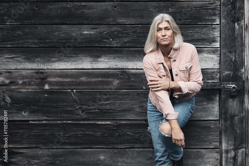 Older woman in her fifties in front of an old wooden wall, dressed in a pink denim jacket, blue jeans and a black T-shirt, in a retro style © RD-Fotografie